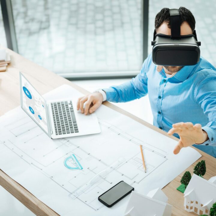 Konzentriert bei der Arbeit. Die Draufsicht auf einen angenehmen jungen Mann, der an einem Laptop arbeitet und eine VR-Brille verwendet, um Konstruktionen zu visualisieren.