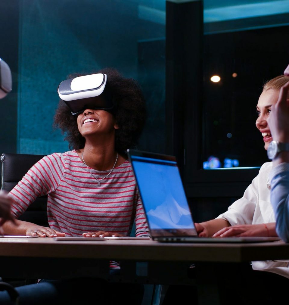 Multikulturelles Geschäftsteam verwendet Virtual-Reality-Headset bei einem nächtlichen Büromeeting. Entwickler treffen sich mit einem Virtual-Reality-Simulator um einen Tisch in einem kreativen Büro.