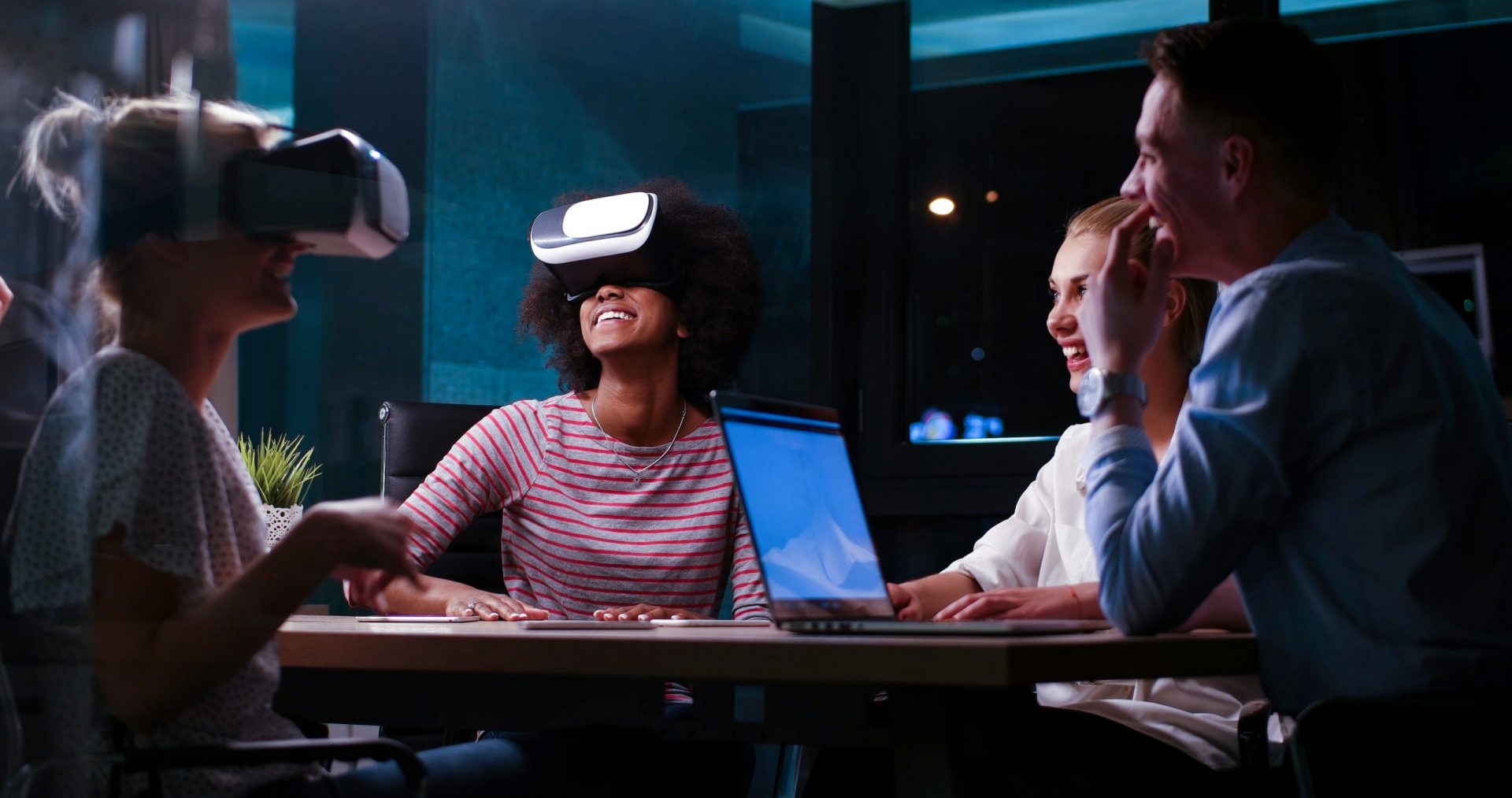 Multikulturelles Geschäftsteam verwendet Virtual-Reality-Headset bei einem nächtlichen Büromeeting. Entwickler treffen sich mit einem Virtual-Reality-Simulator um einen Tisch in einem kreativen Büro.