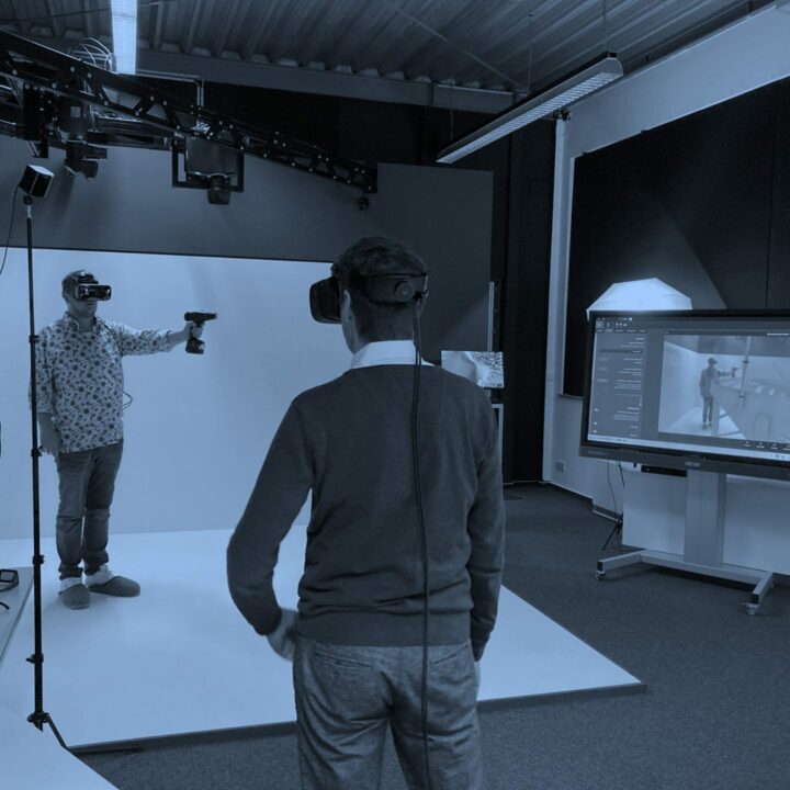 Ein Mann mit VR-Brille und Controller steht vor dem Bildschirm, auf dem seine Abbildung mit einer Maschine zu sehen ist.